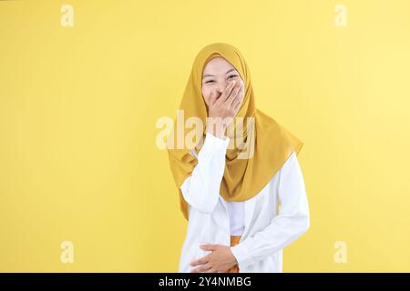 Portrait of Happy Young Asian Woman Giggling in Hijab, Covering Mouth While Trying to Stop Laughing, Isolated on Yellow Background Stock Photo