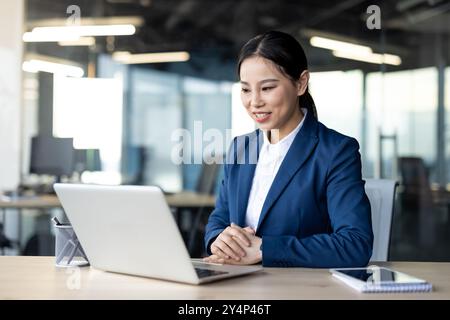 Confident Asian businesswoman wearing blue suit engaging in video call using laptop in modern office. Smiling and focused on digital meeting, showcasing communication and technology Stock Photo
