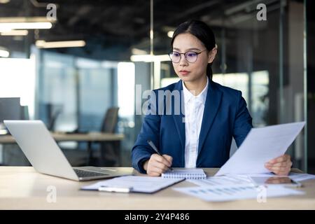 Businesswoman studies financial documents in office. She focuses on paperwork, analyzing data while using laptop. Professional setting with determined investor reviewing reports Stock Photo