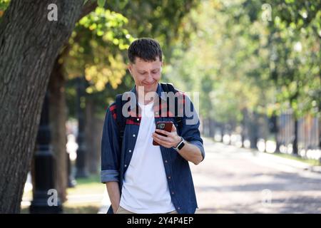 Smiling man with smartphone walking down the street. Using mobile phone in city park at autumn Stock Photo