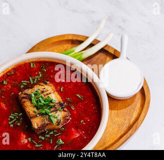 Vibrant bowl of borscht garnished with tender pork, fresh herbs, on a wooden tray with a white background Stock Photo