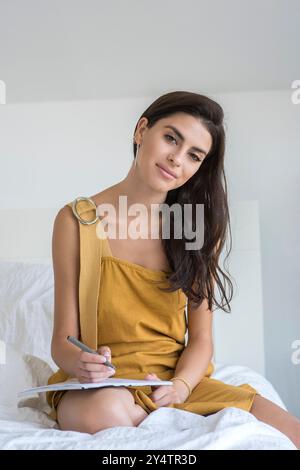 Young woman taking notes in bed. She is a brown dress and has long black hair. Stock Photo
