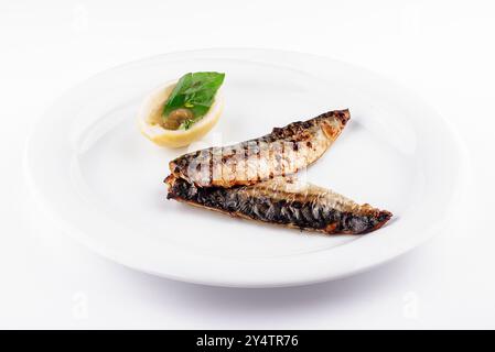 Two grilled mackerel fillets are served on a white plate with a lemon and basil garnish Stock Photo