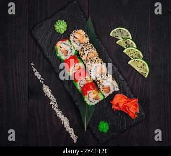 Two types of sushi rolls are laying on a black stone plate, decorated with lime, wasabi and ginger Stock Photo