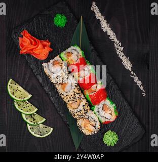 Two types of sushi rolls are laying on a black stone plate, decorated with lime, wasabi and ginger Stock Photo