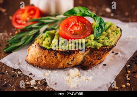 Toasted multigrain bread topped with mashed avocado, a cherry tomato and basil leaf on a wooden table Stock Photo