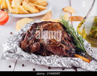 Delicious roasted lamb leg seasoned with rosemary and spices, served on a white wooden table with roasted potato wedges Stock Photo