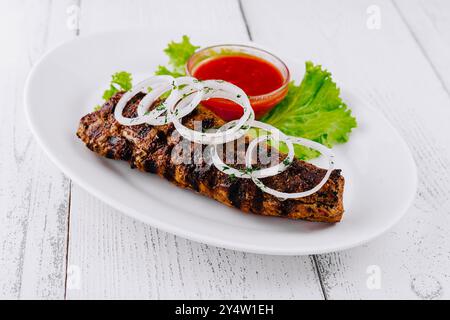 Grilled lula kebab served on a white plate with onion rings, greens, and spicy sauce is laying on a white wooden table Stock Photo