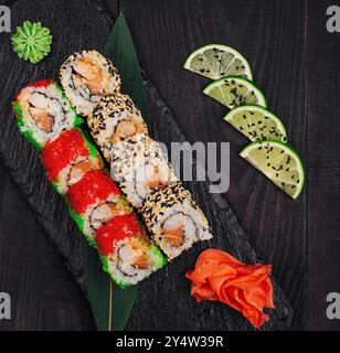 Two types of sushi rolls are laying on a black stone plate, decorated with lime, wasabi and ginger Stock Photo