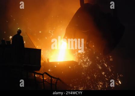 The image captures a dramatic scene inside a foundry or steel mill, showing the process of molten metal being poured from a large ladle into a mold or Stock Photo