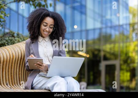 Hispanic businesswoman works outdoors holding notebook and using laptop for online meeting. Shows confidence, professionalism, multitasking in urban environment with glass facade backdrop. Stock Photo
