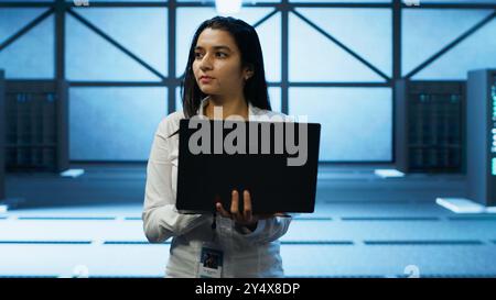 Programmer walking in data center, using notebook to design, implement and manage gear, ensuring optimal performance and reliability. IT professional in server hub coding on laptop, camera A Stock Photo