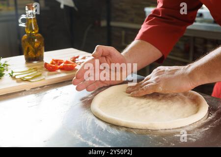 A person shaping pizza dough on a floured surface in a kitchen, with ingredients like tomatoes and pickles in the background. Stock Photo