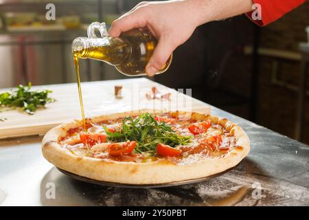 A hand pouring olive oil onto a freshly baked pizza topped with arugula and tomatoes in a kitchen setting. Stock Photo
