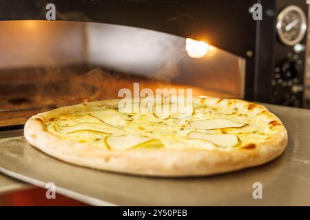 A freshly baked pizza with thin slices of pear on top, placed on a metal tray. The pizza is in a wood-fired oven, with steam rising in the background, Stock Photo