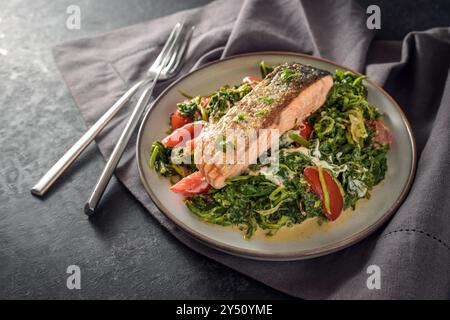 Salmon fillet roasted with a crispy skin on cream spinach vegetables with onions and tomatoes, healthy fish dish with cutlery on a dark gray napkin an Stock Photo