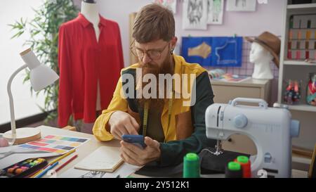 Handsome redhead man with beard using smartphone in tailor shop full of sewing items and mannequin. Stock Photo