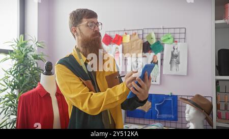Bearded man using smartphone in tailor shop with mannequin and fashion sketches. Stock Photo
