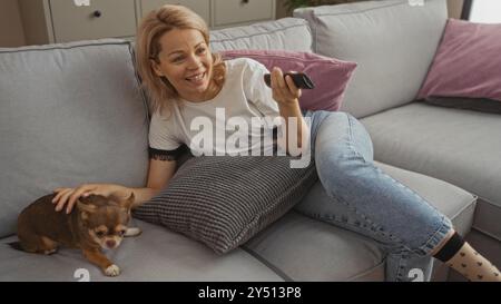 A young blonde woman lounges on her living room couch with her pet chihuahua, smiling and holding a remote, creating a cozy indoor atmosphere. Stock Photo