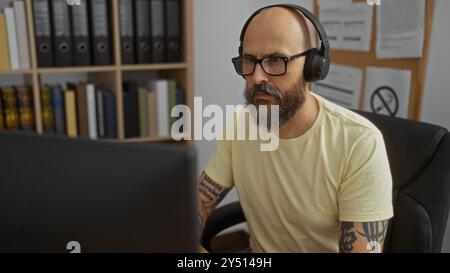 Handsome man with beard wearing headphones working in an office, seated at a desk with shelves and documents in the background, focused on his compute Stock Photo