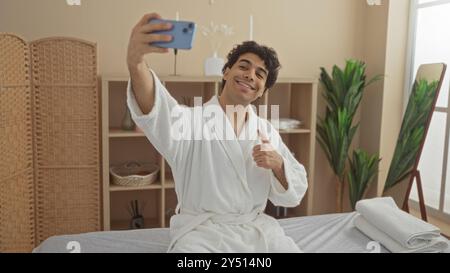 Young man in spa making a thumbs-up gesture while taking a selfie, wearing a white bathrobe in a wellness center Stock Photo