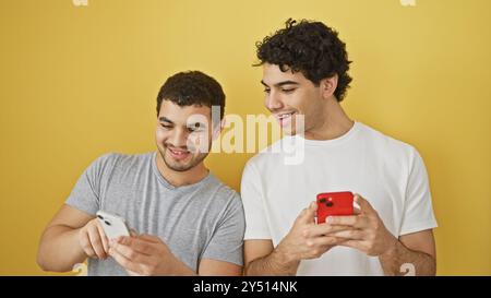 Two hispanic men smiling and using smartphones against a yellow background Stock Photo
