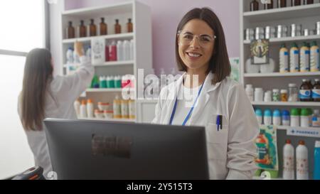 Two women pharmacists working together in a well-organized pharmacy, with one woman smiling at the camera and the other arranging products on the shel Stock Photo