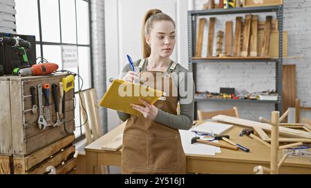 A focused young woman notes details on a clipboard in a well-equipped carpentry workshop. Stock Photo