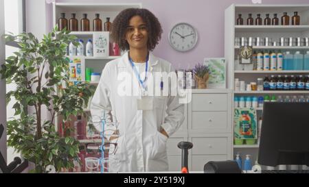 A beautiful young african american woman with curly hair stands confidently in a pharmacy, wearing a white lab coat indoors. Stock Photo