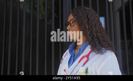 African american female doctor with curly hair stands outdoors in urban setting near a hospital, wearing eyeglasses and a stethoscope. Stock Photo