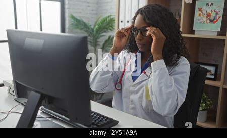 Young african american woman with curly hair in a white coat and glasses, working at a computer in a clinic's indoor office. Stock Photo