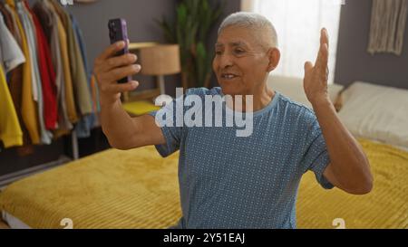 A mature hispanic man with grey hair is making a videollam from his bedroom at home. Stock Photo