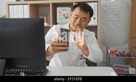 A young, handsome asian man in a white coat waves while video calling on a smartphone in a clinic office equipped with shelves and office supplies. Stock Photo