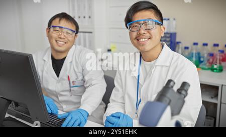 Two smiling men wearing lab coats and safety glasses working together at a computer in a laboratory setting. Stock Photo