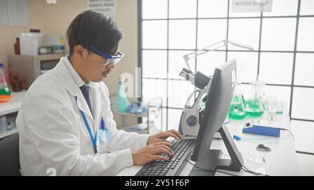 A young asian man in a lab coat working on a computer in a bright laboratory setting. Stock Photo