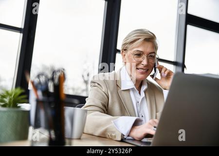 An adult woman dressed elegantly engages in a phone conversation while working on her laptop. Stock Photo