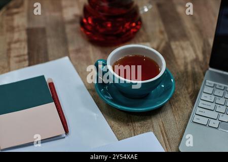 High angle shot of turquoise ceramic cup of black tea on wooden table with laptop and notes Stock Photo