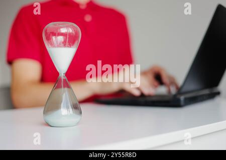 Hourglass with flowing sand on wooden table. Man typing on a laptop keyboard indoors. Concept of time management and deadline. Stock Photo