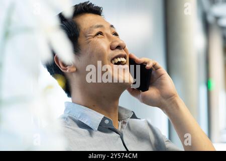 Talking on smartphone, asian man smiling and enjoying conversation in office setting Stock Photo