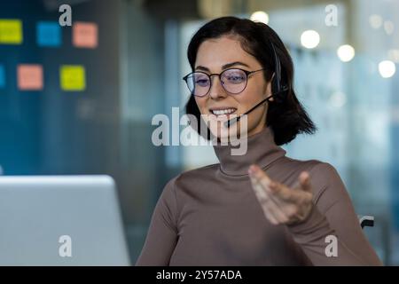 Businesswoman using laptop wearing headset for online meeting. Smiling while gesturing during virtual conversation in modern office setting. Professional setting with focus on customer service Stock Photo