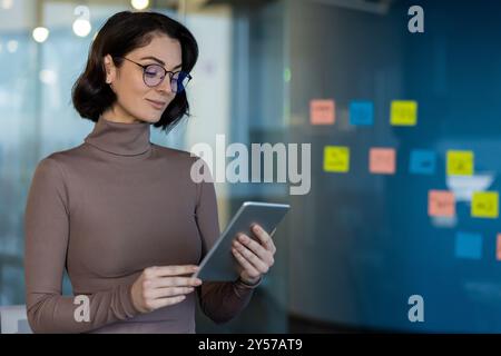 Businesswoman holds tablet engaging in work activities in office. Confident professional using technology for planning and organization, wearing glasses and casual attire. Stock Photo