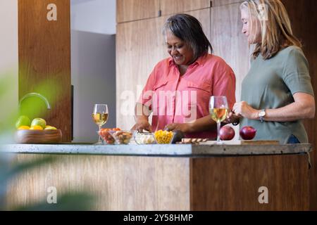 At home, preparing meal, two senior multiracial female friends enjoying wine and cooking together Stock Photo