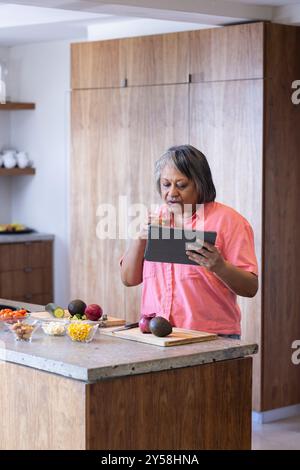 At home, preparing meal, senior woman using tablet and drinking wine in kitchen Stock Photo