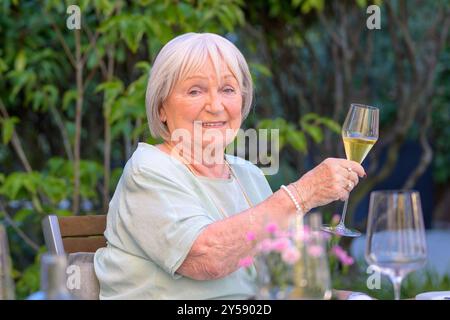 A cheerful elderly woman holding up a champagne glass in a toast while seated at an outdoor table surrounded by greenery, celebrating in a relaxed set Stock Photo