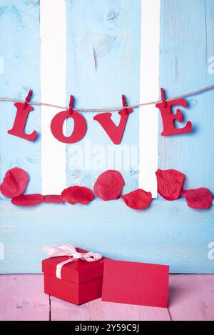 Cute red gift box with a blank message card near it and the word love written in red paper letters tied to a string, on a blue wooden fence Stock Photo
