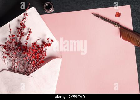 Romantic frame with an envelope full of red flowers, on a blank paper sheet, near an ink bottle and an old feather pen, on a black wooden table Stock Photo