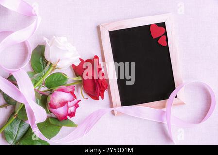Lovely bouquet of multicolored roses, tied with a pink ribbon, near a blank chalkboard decorated with two red hearts, on a pink background Stock Photo
