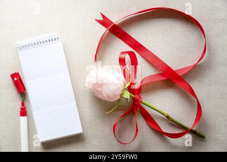 Pink rose tied with a red ribbon in shape of an 8 and an open blank spiral notebook with stripes and a red marker pen near it, on a vintage fabric Stock Photo