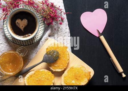 Romantic breakfast with flowers, heart shaped sticky note, bread with jam on a trencher and a cup of coffee with a foam heart, on a black table Stock Photo