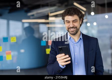 Businessman wearing suit using smartphone in modern office. Man smiling, engaging with phone for work communication or tasks. Office environment with note-covered glass board in background. Stock Photo